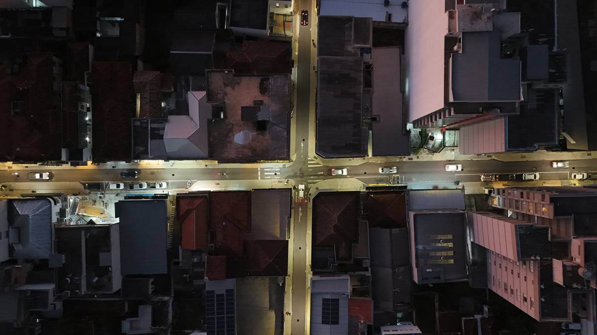 Vista aérea noturna de um cruzamento urbano iluminado, com as ruas em cruz acesas entre os telhados