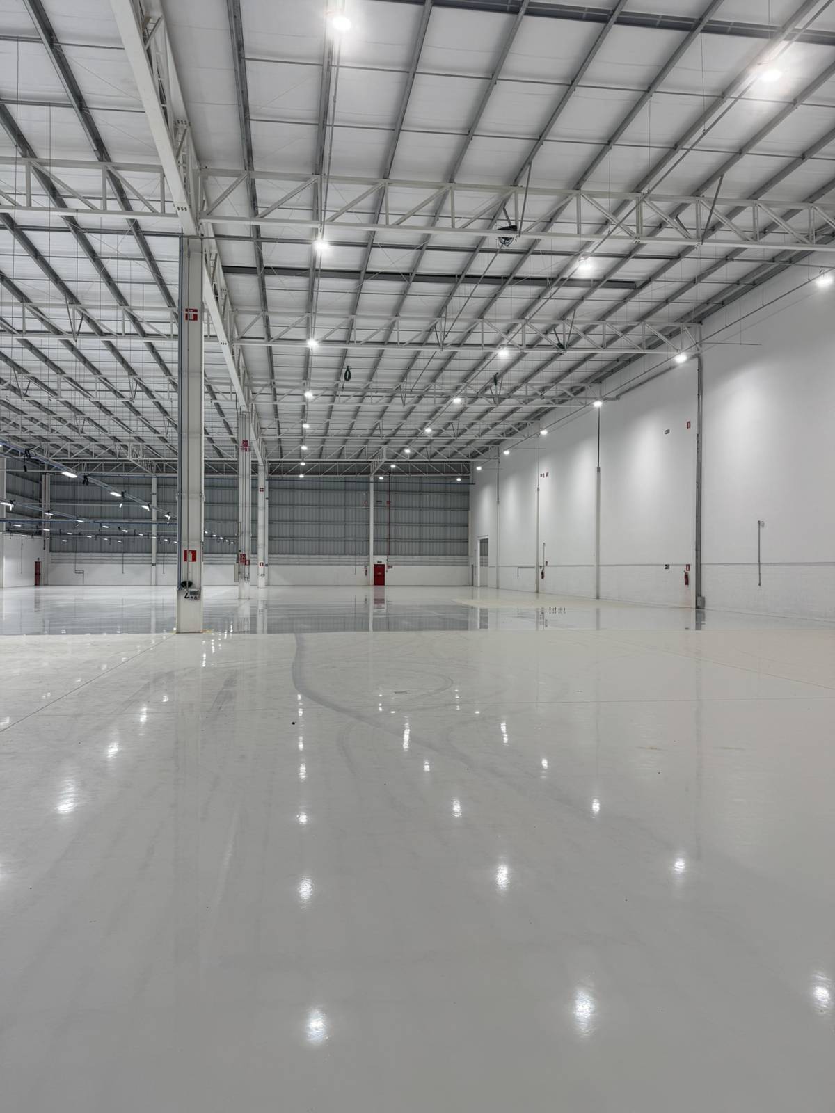 Galpão industrial com luminárias industriais suspensas acesas, refletidas no piso de concreto polido