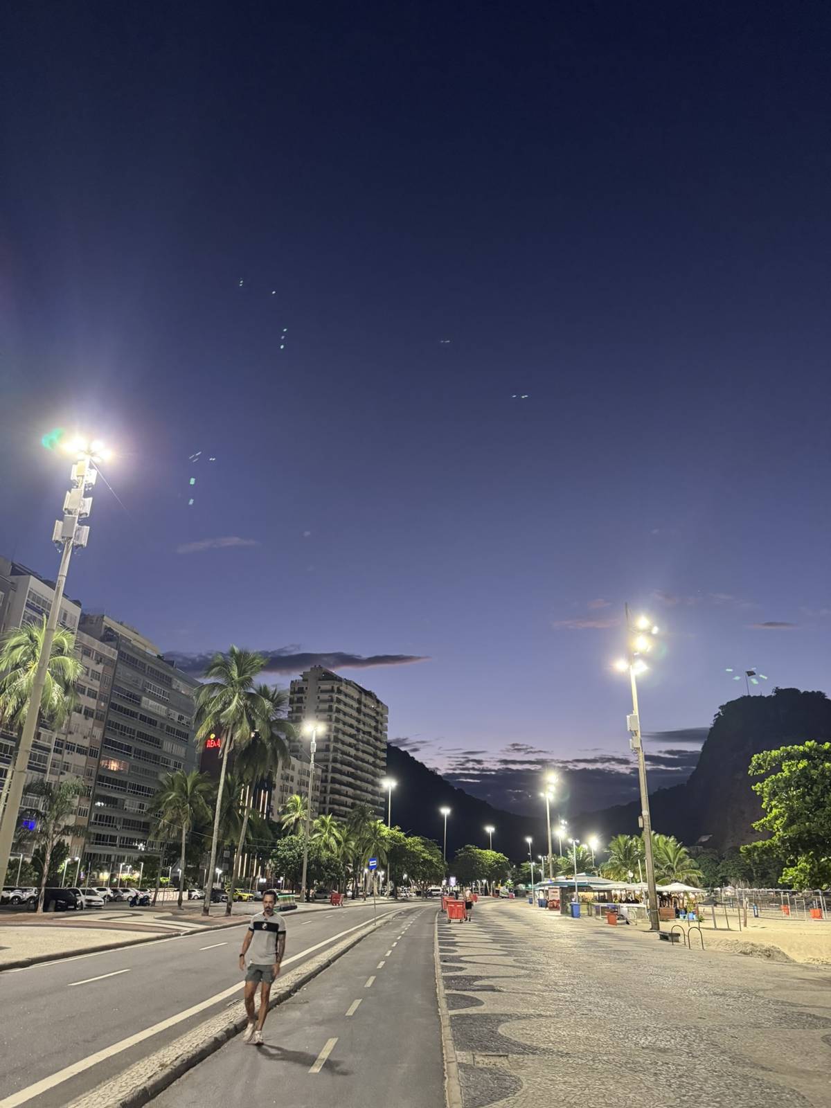 Orla urbana ao anoitecer com postes altos iluminando o passeio, a ciclovia e as palmeiras à beira-mar