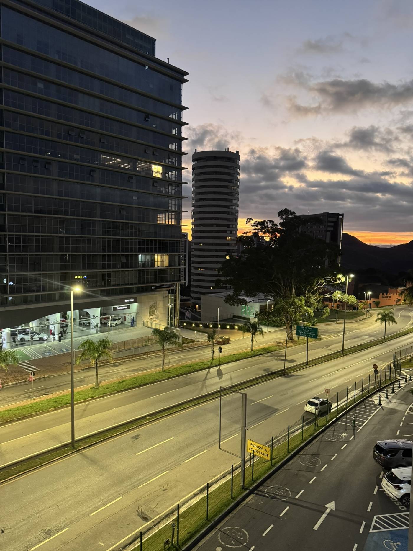 Avenida de múltiplas faixas ao entardecer, com a iluminação pública já acesa sobre as pistas
