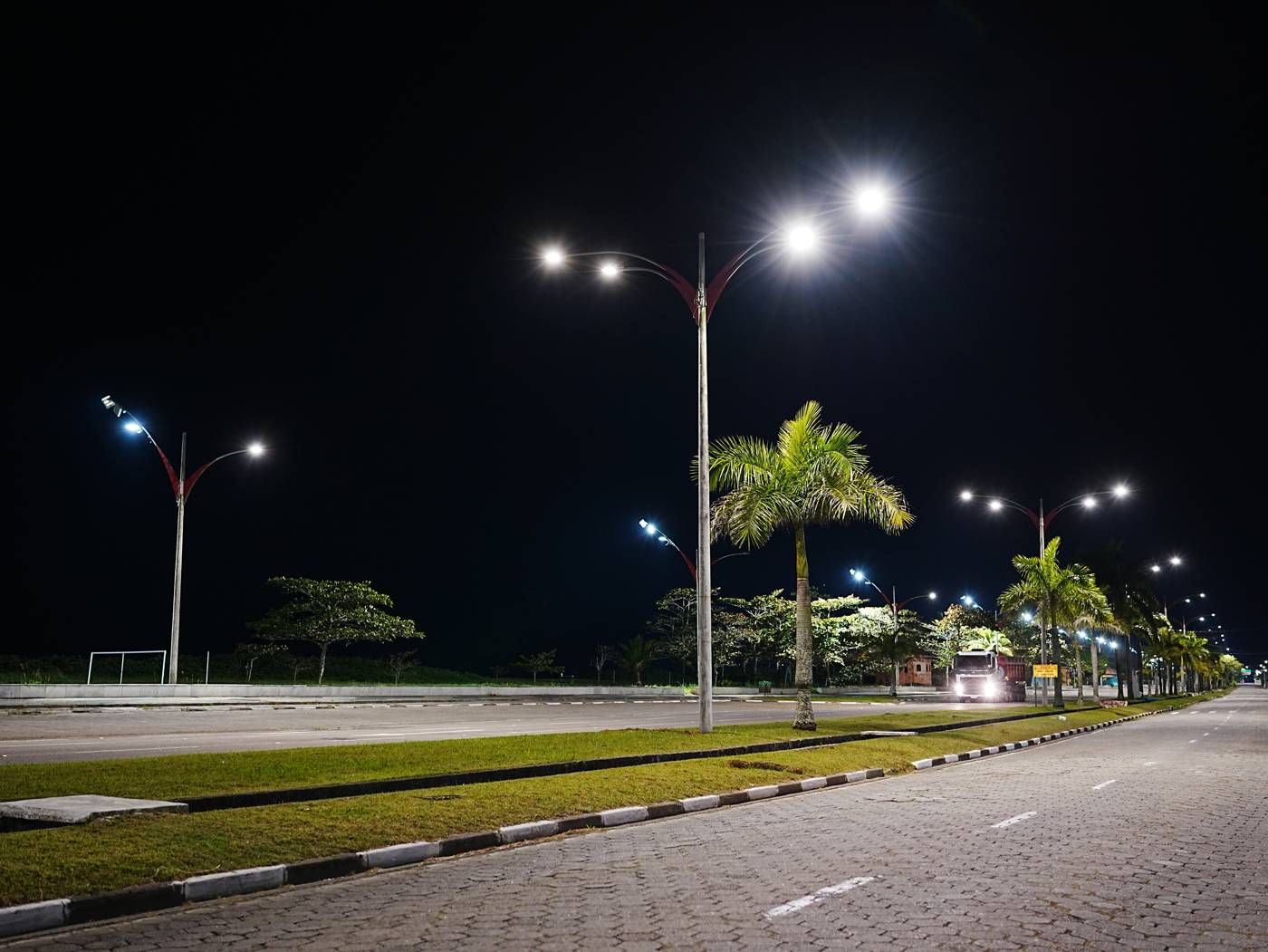 Avenida urbana à noite com postes de iluminação pública acesos, ladeada por palmeiras