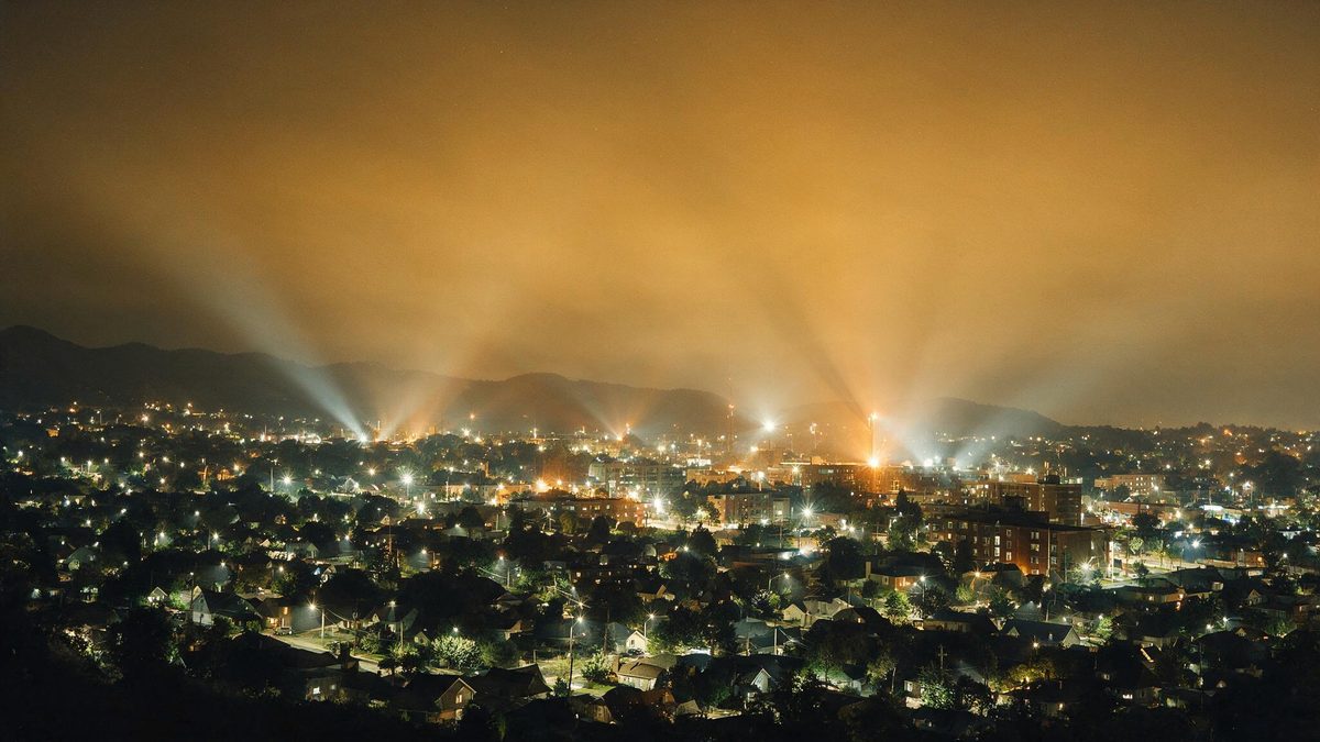 Postes de rua espalhando luz para o céu numa ladeira, com igreja iluminada ao fundo