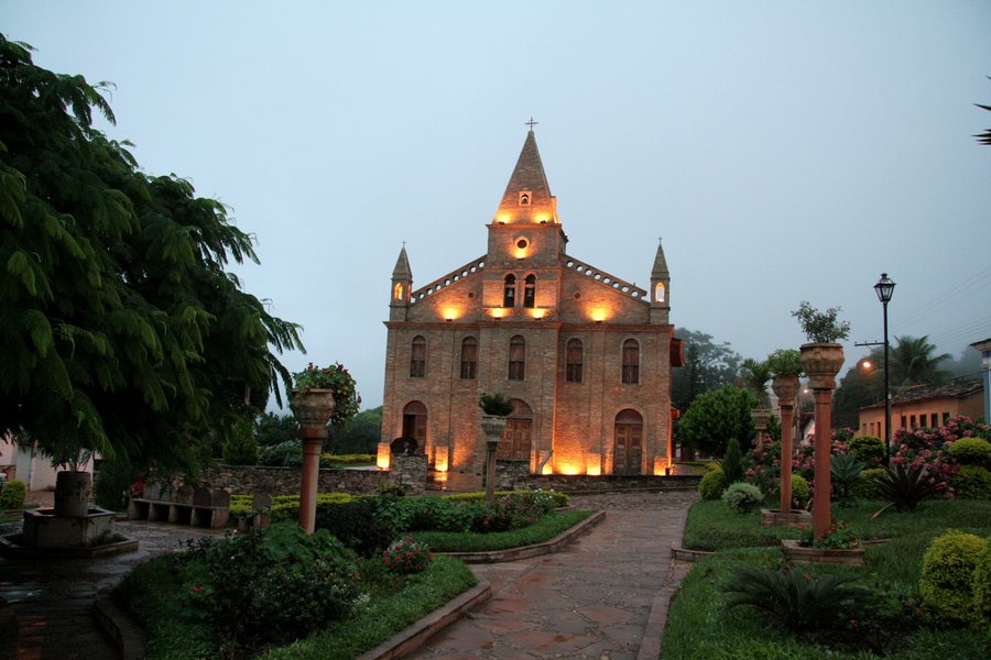Igreja iluminada em âmbar ao fundo de uma praça ajardinada, no fim da tarde